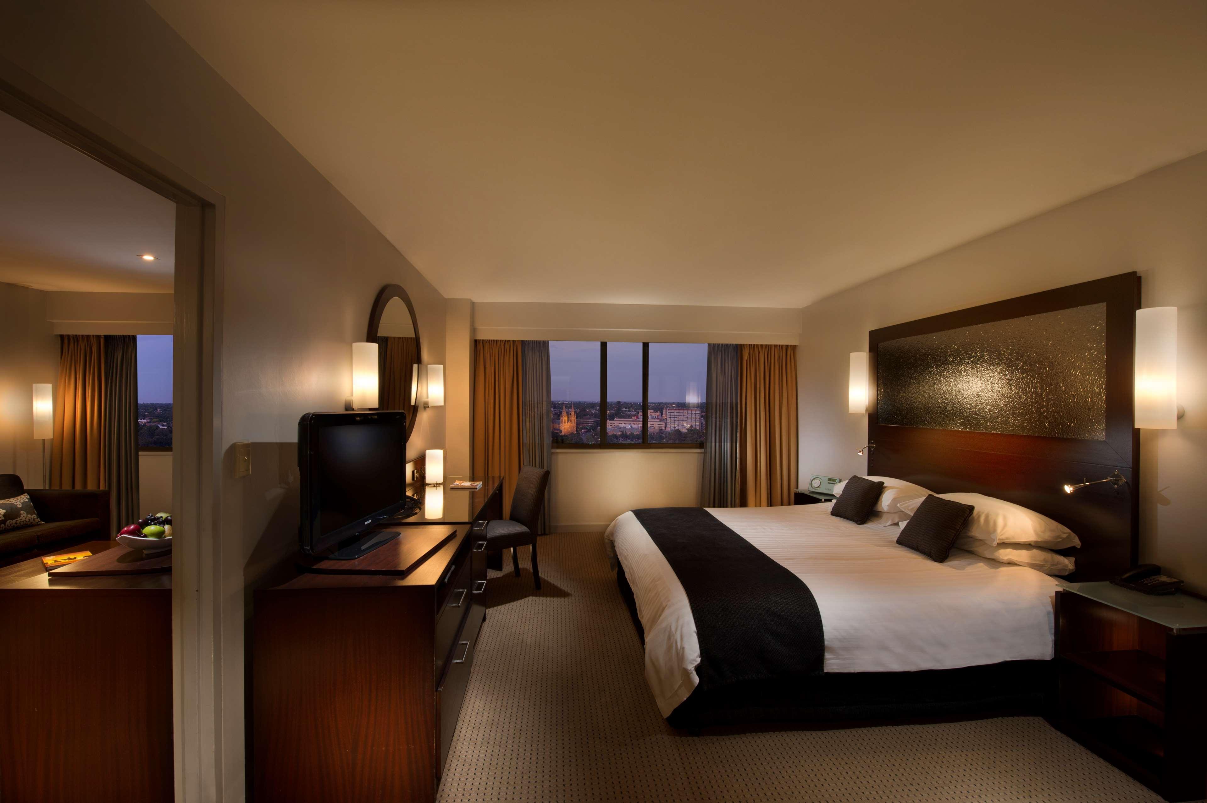 Stamford Plaza Adelaide Hotel Luaran gambar The photo features a modern hotel room that includes a large bed with dark and light bedding, complemented by two pillows. The room is well-lit with warm lighting and has a contemporary aesthetic. To one side, there is a wooden dresser with a flat-sc